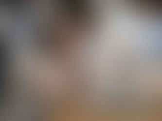 Eine Frau sitzt an einem Schreibtisch im Büro und hält sich mit beiden Händen den Kopf. Kolleg:innen arbeiten unscharf im Hintergrund.