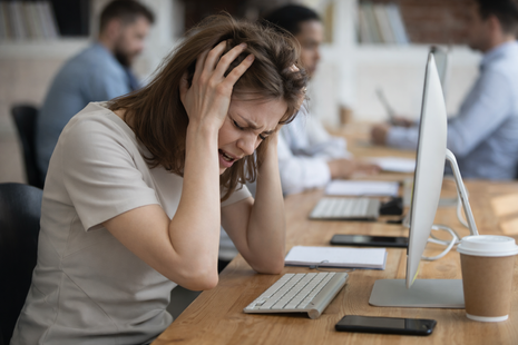 Eine Frau sitzt an einem Schreibtisch im Büro und hält sich mit beiden Händen den Kopf. Kolleg:innen arbeiten unscharf im Hintergrund.