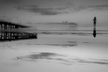 Eine einzelne Person steht weit entfernt am Strand, alles wirkt ruhig und leer, während sich Himmel und Wasser fast spiegeln und eine distanzierte, gefühllose Stimmung entsteht.