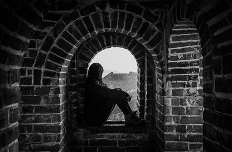 Eine Person sitzt in einer dunklen Steinöffnung und blickt nach draussen in eine helle Landschaft, still und in sich zurückgezogen.