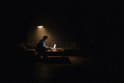 Dunkles Wohnzimmer, nur ein enger Lichtkegel erhellt einen Tisch mit Laptop. Eine Person sitzt genau in diesem Licht, der restliche Raum verschwindet vollständig im Schatten. Die Szene wirkt still, fokussiert und isoliert