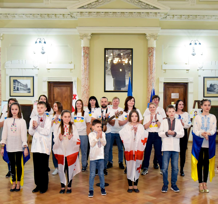 A group of 19 children and adults wearing Ukrainian national costumes stand performing national anthems of Georgia and Ukraine