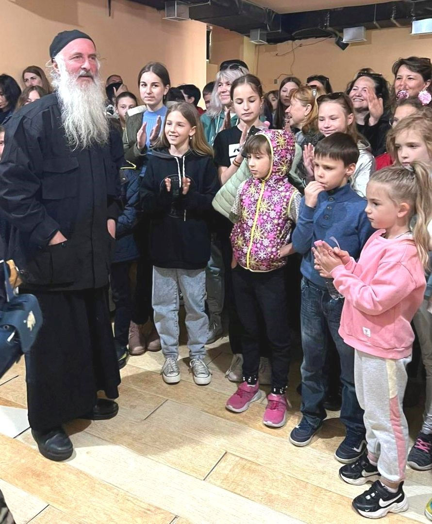 Metropolitan Isaia, a bearded man in traditional monk's robes speaks enthusiastically to a group of children