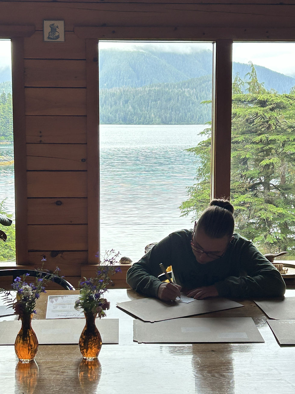Woman writing at a table beside a window overlooking the water