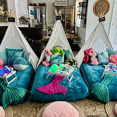 picture of a living room for a child's birthday party. the furniture has been moved and 3 teepees are placed in the living room with mattresses. all of the teepees are decorated with multi-colored string lights, blue plush blankets, as well as colorful stuffed animals such as a pufferfish, octopus, turtle, seahorse. On top of each mattress is a tray that has a whale shaped mirror, a flowery lei, and a light up globe.