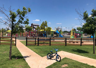 A lone bicycle sits on a sidewalk that interrupts the beautiful green grass of a park. In the background, under a clear blue sky, jungle gyms and other playground activities await the youth for adventures at Wild Horse Park.