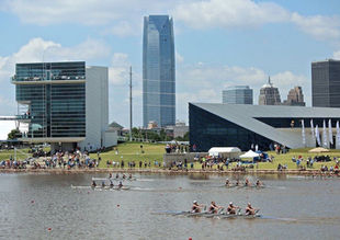 A vibrant photo of the Boathouse District in Oklahoma City, showcasing sleek, modern boathouses along the Oklahoma River. People are kayaking and rowing on the water, while others enjoy the riverside trails and green spaces under a bright blue sky.