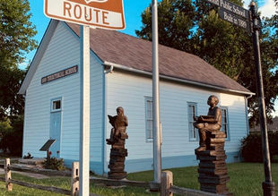 The exterior of the historic white one-room school house. The scene also includes two bronze sculptures of children sitting atop a stack of books while reading, a historic route 66 street sign, a US flag, and a short wooden fence that surrounds the area.