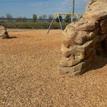 A wide slender tan colored faux rock formation sits in an area of a park covered with tan pebbles.