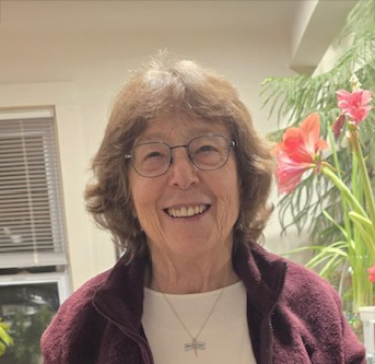 A headshot photo of a smiling, older white woman with chin-length, brown hair and glasses. She is wearing a maroon fleece jacket over a white shirt and a silver dragonfly necklace. There are a pair of large coral tropical flowers and greenery just behind her to the right, with a white wall and window in the background.
