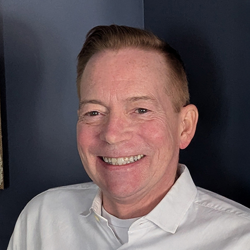 A headshot photo of a smiling, middle-aged white man with short, redish-brown hair. He is wearing a white collared shirt and the background is dark gray.