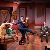 5 actors stand onstage in a drawing room. One actor is being held by two others in a compromising position,