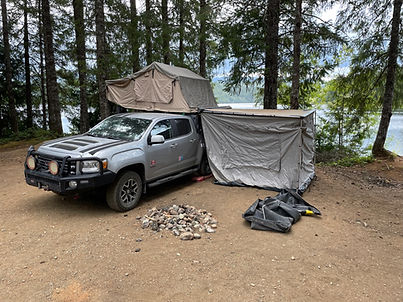 A picture of a GMC Canyone with a roof top tent, and an awning with a room, overlooking a mountain lake,