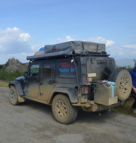A Jeep Wrangler JK with a roof tent, an awning, some spare fuel cans, covered in mud with "Prudhoe Bay or Bust" written on the window.