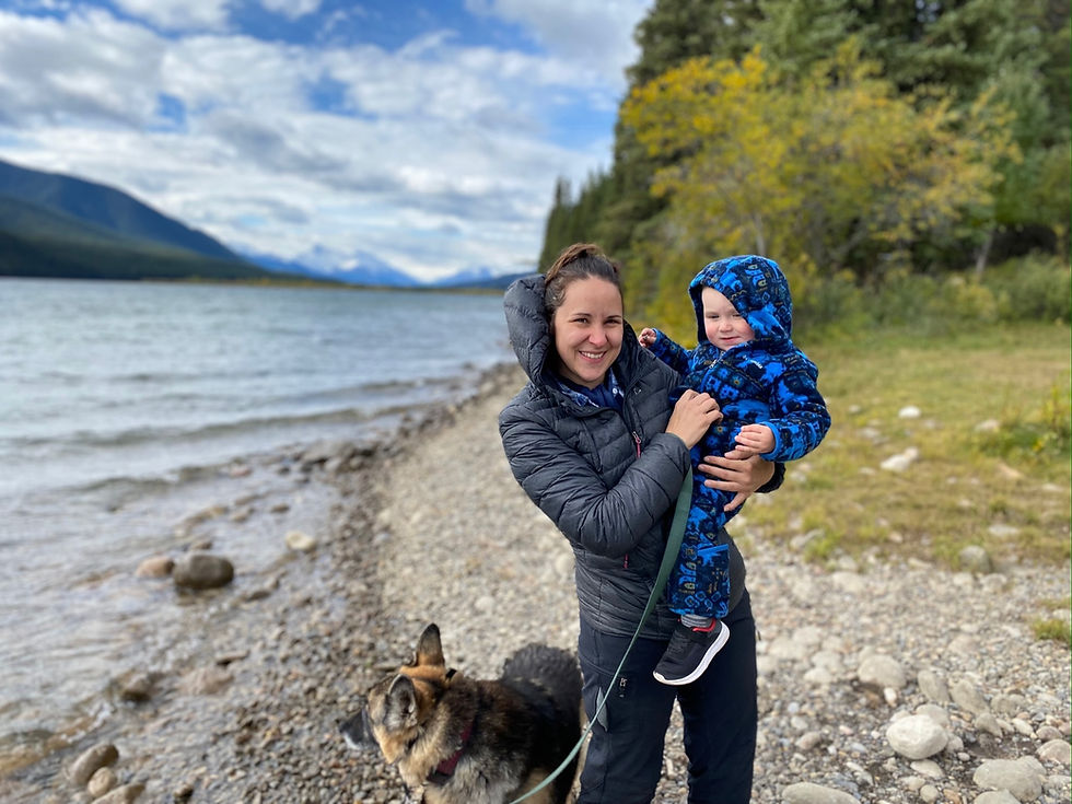 A woman, Teryn, standing on the shore of a mountain lake holding a baby. They are bundled up in warm clothes but it appears to be fall or spring. There's also a German Shepherd attached to a leash in her hand.