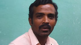 Manjish, an Indian trans man who is also intersex, standing outdoors. He is dark-skinned with short, black hair and black facial hair. He is wearing a collared pink shirt and is speaking, looking directly at the viewer. The background of the photo is a bright green wall.