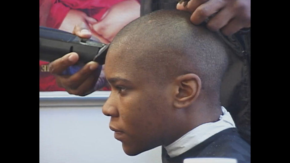 Octavio, a young trans man in the documentary The Aggressives, sitting in a barber shop and looking ahead with a neutral or angry expression. He is dark-skinned with a buzzcut.