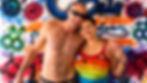 Julian Melson, an American trans man, standing in front of a graffiti-covered wall with an American woman named Theresa Campos. Both are light-skinned and smiling happily. He has his arm around her shoulders, and she is holding onto his arm in an affectionate way. He is wearing shorts. She is wearing a rainbow shirt with the words "I love my trans friend," written on it. He has balding, grey hair, and is wearing dark sunglasses. She has short, black hair, and is wearing makeup. The graffiti includes gay, lesbian, transgender, and other pride symbols.