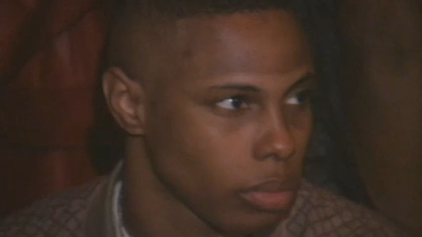 Marquise Vilsón, a Black trans man in the documentary The Aggressives. He is sitting indoors, looking into the distance with a neutral expression. He has short, black hair, and is wearing a brown coat over a white shirt.