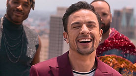 Luca Scarpelli, a Brazilian trans man, in the Brazilian Queer Eye show. He is outdoors, smiling happily, eyes closed. He has short, dark hair and dark facial hair. He is wearing a red jacket over a white t-shirt. His co-hosts are dancing around him, partly visible in the background, with a city visible in the blurred background.
