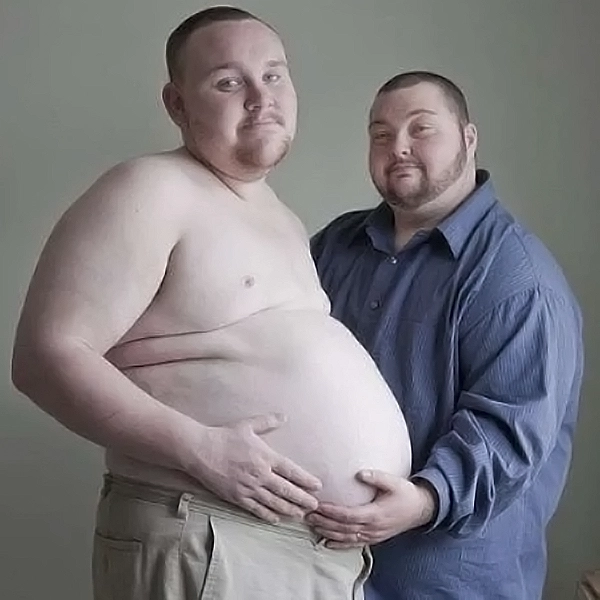 Two trans men standing together. One is holding his husband's bare pregnant belly