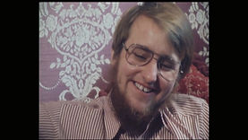 Steve, an English trans man, in the 1980 British documentary What Am I? He is a light-skinned man with short, light brown hair, with a bushy, light brown beard. He is laughing happily. He is wearing wire-rimmed glasses and a red-and-white chequered, collared shirt. Patterned pink wallpaper can be seen behind him.