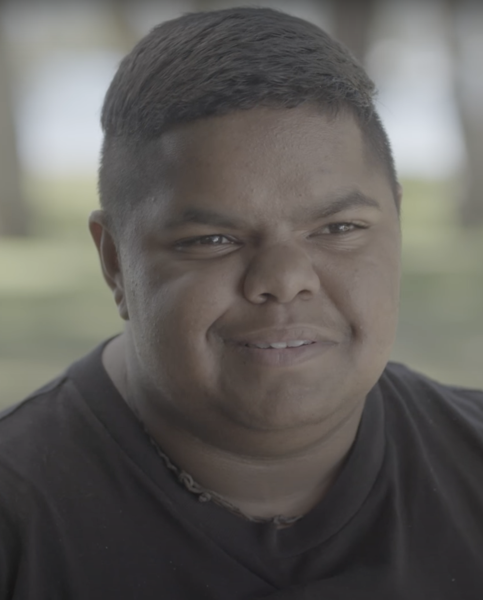 A trans man grinning. He is wearing a black shirt and has dark skin.