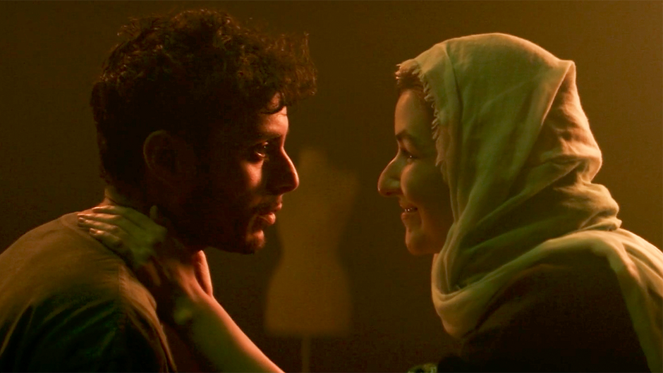 Adam Kashmiry, an Egyptian-Scottish trans man in the film Adam. He is standing indoors. A lesbian woman named Amira, played by Rehanna MacDonald, is standing in front of him, hands on his shoulders, looking into his eyes and smiling. She is wearing a head scarf and has long, dark hair. He has short, dark hair and a neutral expression.