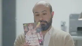 Ahmad, a trans male character played by Iranian-American trans man Aden Hakimi, sitting indoors and reading a sexual health pamphlet about sex with trans men