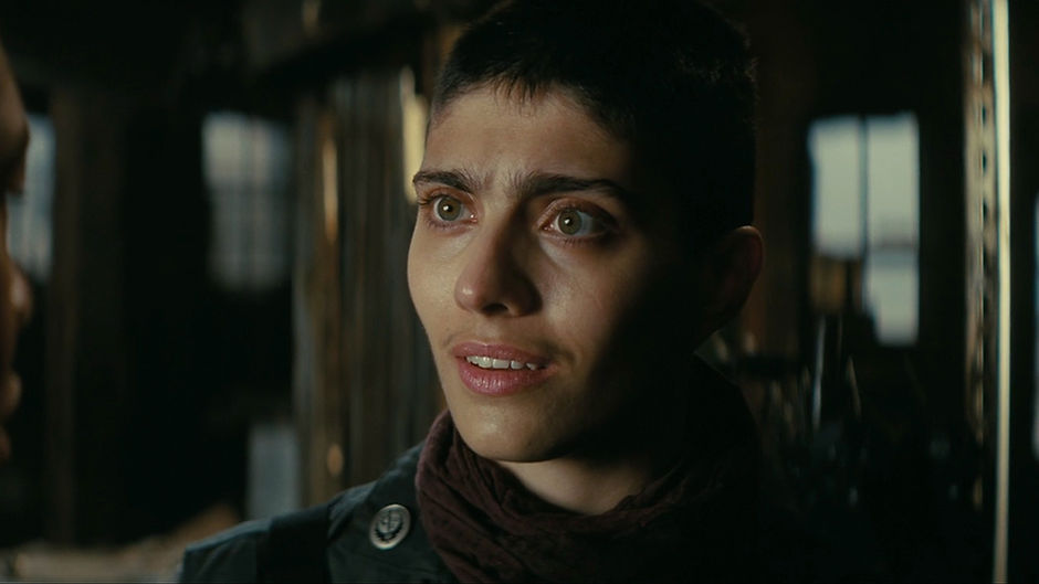 Dane, a non-binary person played by Indian-English trans actor Xelia Mendes-Jones, in the show Fallout. They are standing indoors, looking into the distance with a sincere or earnest expression. They have light brown skin, short black hair, hazel eyes, and a faint black moustache. They are wearing a dark red turtleneck shirt and a black vest. The interior of a military facility is visible in the blurred background.