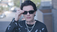 Jesse Sullivan, a trans man, in the documentary My Transparent Life. He is walking outdoors and adjusting his dark sunglasses. He has short, black hair, light skin, and a tattooed neck. He is wearing a patterned black sweater and silver chain necklaces.