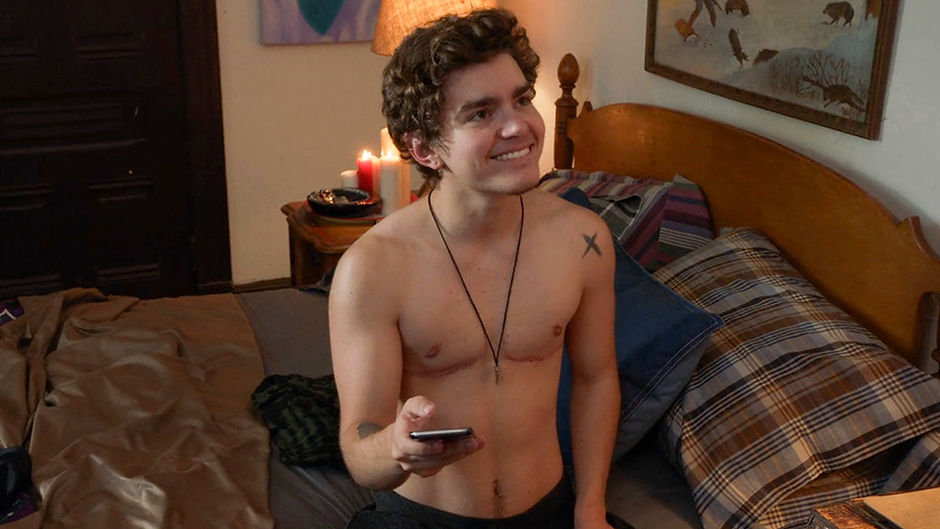 Trevor, a trans man played by American trans male actor Elliot Fletcher, in the show Shameless. He is sitting on a bed, holding a remote and grinning. He has short, dark curly hair and visible top surgery scars. He is wearing dark trousers. He has a cross tattoo on his shoulder.
