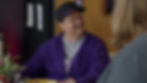 Fred Rococo, a trans man played by American trans actor Murray Hill, in the show Somebody Somewhere. He is light-skinned with dark, short hair, wearing a purple hoodie over a grey shirt. He is sitting at a table and looking up at someone, smiling. Someone is visible in the blurred foreground, watching the interaction.