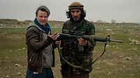 Jordan Bryon, a middle-aged, light-skinned, grey-haired Australian trans male journalist in the documentary Transition. He is standing outdoors in a field, wearing a green coat over a grey hoodie over blue perahan tunban. He is standing beside a darker-skinned Afghan man who is wearing a camouflage uniform and a light brown pakol hat. The man is holding a large gun, and Jordan is gesturing, looking toward the viewer with a neutral expression.