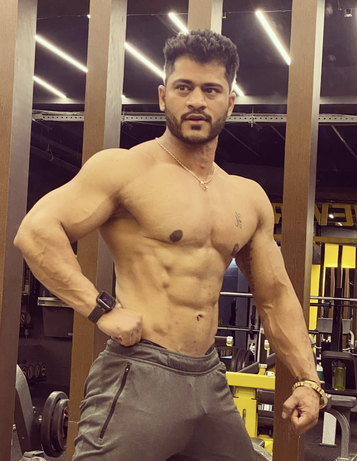 An Indian trans man posing while wearing grey shorts. He is muscular, has trimmed facial hair, and faint top surgery scars. He has a visible crotch bulge.