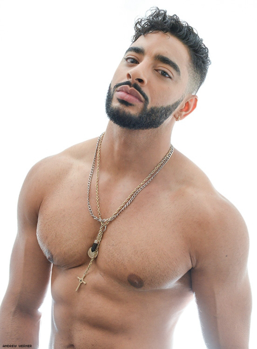 A very muscular man with a thick, dark goatee and a partly-shaved head, wearing a necklace against his bare chest.