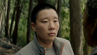 Pin-Lee, a non-binary lawyer played by American-Chinese non-binary actor Sabrina Wu, in the show Murderbot. They are a lighter-skinned person with short, black hair and dark brown eyes. They are wearing small, subtle hoop earrings, a grey jacket, and a grey t-shirt. They are looking into the distance with a neutral expression. A daytime forest can be seen in the blurred background.