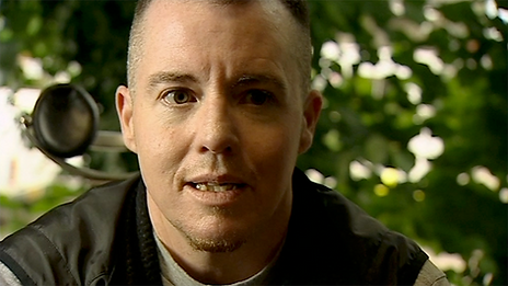A light-skinned intersex non-binary person sitting indoors, looking to the side as they speak directly to the viewer. They have short, dark brown hair and faint brown facial hair. They are wearing a black vest over a light green shirt. Trees can be seen in the blurred background, through a window.