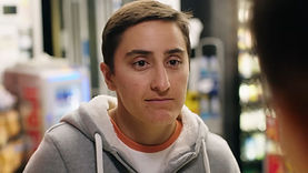 Kirby, a trans man played by trans actor Eisa Al-Shamma, in the short film (In)convenience. He is standing indoors in a convenience store, smiling slightly. He has light brown skin, dark brown eyes, and short brown hair. He is wearing a grey hoodie over a white shirt.