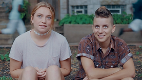 Cyd and Katie, two characters in the film Princess Cyd, sitting together outdoors. Katie is a non-binary person with light skin and partly-shaved brown hair, wearing a patterned brown shirt. Cyd is a woman with long blonde hair, wearing a white shirt with a white neck choker. The pair are looking up with relaxed and neutral expressions.