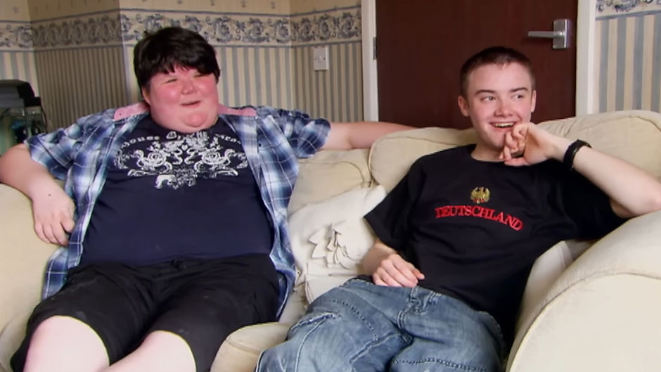 Two light-skinned English trans men sitting on a couch together, grinning widely. Both have short, dark hair. The man on the left is wearing a blue flannel shirt over a blue patterned t-shirt, and black shorts. The man on the right is wearing a black t-shirt with text on it, and blue jeans.