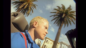 A blond trans man sitting in a car, with palm trees and a blue sky visible in the distance