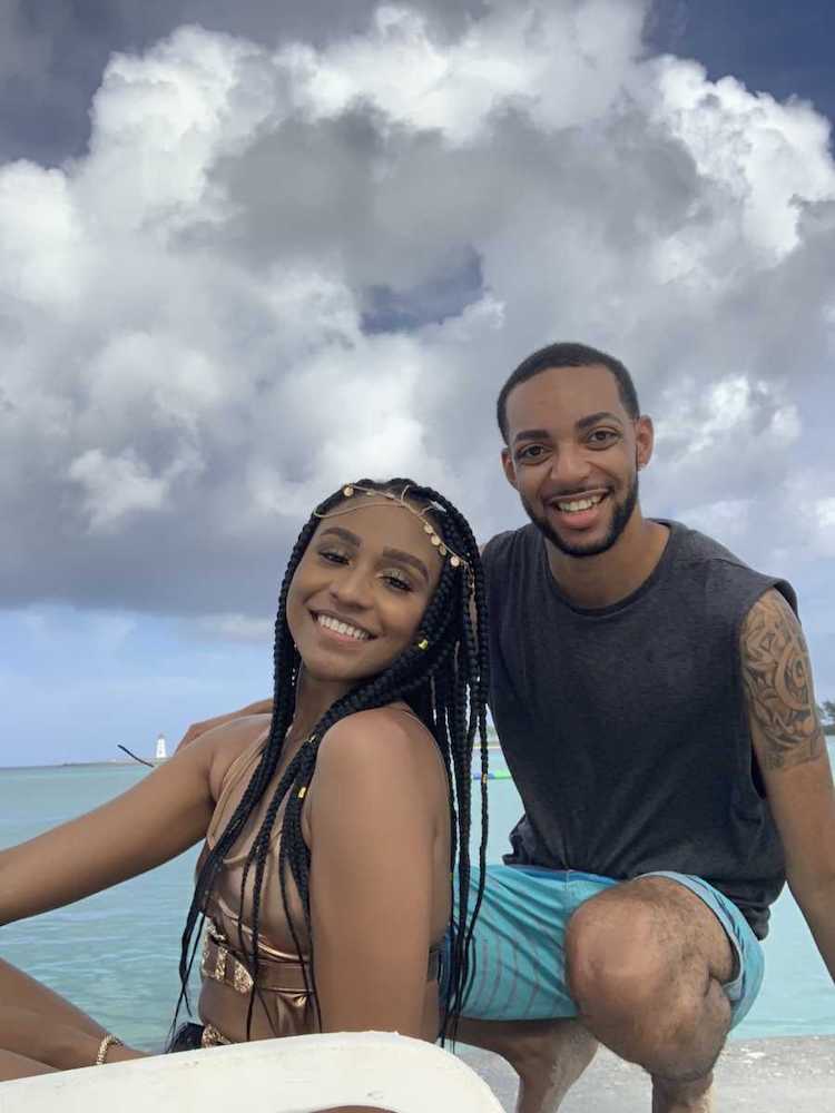 A man and a woman sitting on a beach together. The woman is wearing a rose gold bikini and has her hair braided. The man is wearing a grey tank top and swim shorts.