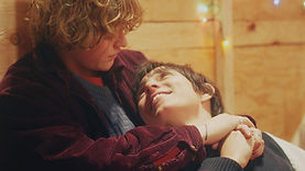 Kieran Dearg, a trans man played by American non-binary actor Izzi Rojas, lying with his head in the lap of Egan Keith, a young man played by American non-binary actor Aidan Dick. Kieran is light-skinned with short, black hair. Egan is light-skinned with short, wavy blond hair.