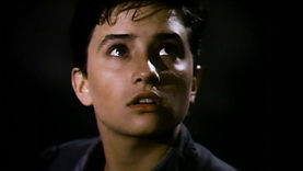 Willy, a trans teen played by American actress Pamela Adlon in the film Willy/Milly, standing outdoors at night. He is looking up with a neutral or interested expression. He is light-skinned with short, wavy black hair. He is wearing a grey flannel shirt.