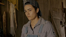 Hank, a trans man played by Thai-American actor Adrian Hill, in the short film Queer and Southern God. He is a brown-skinned man with short, black hair, wearing a patterned cap backwards on his head. He has dark brown eyes and a serious or neutral expression. He is wearing denim overalls over a collared purple shirt. He is looking into the distance.