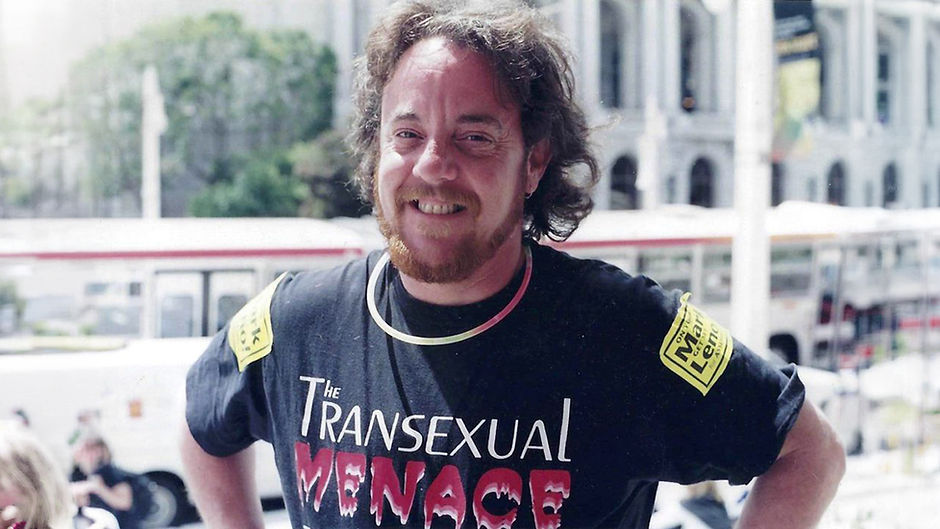 Two-Spirit trans man Marcus Arana standing outdoors, smiling happily. He is light-skinned with shoulder-length, wavy light brown hair, a light brown beard, and dark eyes. He is wearing a black shirt with the words "TRANSEXUAL MENACE" printed on it. He is standing with his hands on his hips. A train, buildings, and trees can be seen in the blurred background.