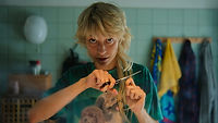 Tosiek, a trans man played by Polish non-binary actor Alin Szewczyk, in the film Fanfic. He is standing in a bathroom, cutting his long blond hair with a pair of scissors. He is wearing a green t-shirt with an illustration on it. Green tiles and clutter can be seen in the blurred background.