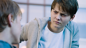 Kim, a teenage trans boy played by Swedish actress Tuva Jagell, in the film Girls Lost. He is light-skinned with short, brown hair and blue eyes. He is sitting outdoors, looking at a boy in the blurred foreground. He is wearing a white t-shirt and a grey hoodie.