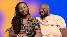 Ja'Mel Ashely Ware, a Black trans man sitting indoors, wearing a peach yellow t-shirt. He is bald, with a black beard. He is sitting next to his partner, a feminine Black gay man with long, braided black hair, wearing a multicoloured patterned t-shirt. The background of the image is an orange and purple gradient.